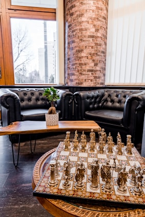 A luxurious interior features two black leather tufted sofas arranged around a wooden coffee table, topped with a small potted plant. In the foreground, an ornate chess set with metal pieces is displayed on a patterned board. The background includes a large window with a view of a cityscape and a prominent brick column.