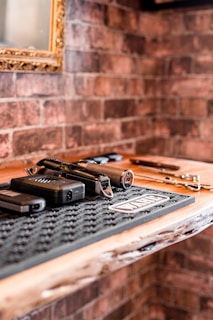 A wooden table displays a handgun along with its magazine laying on a textured mat. The background features a brick wall with a framed mirror.