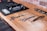 Three barber tools including scissors and a straight razor are laid out on a wooden surface. In the background, an organized station holds clippers and other grooming equipment, contrasted by an exposed brick wall.