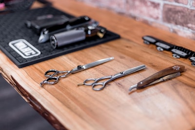 Three barber tools including scissors and a straight razor are laid out on a wooden surface. In the background, an organized station holds clippers and other grooming equipment, contrasted by an exposed brick wall.