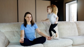A woman is sitting cross-legged on a sofa with eyes closed, appearing to meditate. Next to her, a young girl is energetically jumping on the sofa with a playful expression.