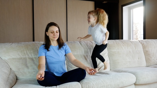 A woman is sitting cross-legged on a sofa with eyes closed, appearing to meditate. Next to her, a young girl is energetically jumping on the sofa with a playful expression.