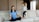 A woman is sitting cross-legged on a sofa with eyes closed, appearing to meditate. Next to her, a young girl is energetically jumping on the sofa with a playful expression.