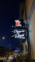 A city street at night featuring illuminated signs for a pizzeria named Pizzeria Rossi, established in 1996. The shopfront includes a black sign with white script and a red and yellow crown emblem above. Streetlights are on, and a few cars and pedestrians can be seen.