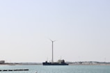 A large wind turbine stands near the shore with a ship positioned parallel to it in the water. The background shows a clear sky and a distant shoreline with some buildings and vegetation.