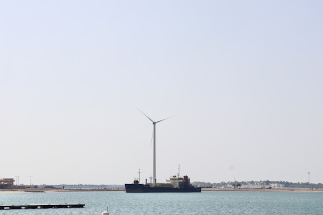 A large wind turbine stands near the shore with a ship positioned parallel to it in the water. The background shows a clear sky and a distant shoreline with some buildings and vegetation.
