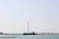 A large wind turbine stands near the shore with a ship positioned parallel to it in the water. The background shows a clear sky and a distant shoreline with some buildings and vegetation.
