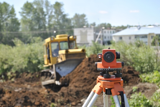 A construction scene featuring a bright orange survey tripod in the foreground, with a blurred yellow bulldozer moving dirt in the background. The scene has green trees and shrubs on the left side and some buildings partially visible on the right side under a clear blue sky.