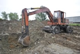 An old, rusty excavator is parked on rough terrain, surrounded by piles of dirt and gravel. The excavator appears to be in working condition with visible hydraulic components. It is set against a backdrop of trees and industrial buildings.