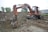 An old, rusty excavator is parked on rough terrain, surrounded by piles of dirt and gravel. The excavator appears to be in working condition with visible hydraulic components. It is set against a backdrop of trees and industrial buildings.