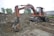 An old, rusty excavator is parked on rough terrain, surrounded by piles of dirt and gravel. The excavator appears to be in working condition with visible hydraulic components. It is set against a backdrop of trees and industrial buildings.