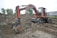 An old, rusty excavator is parked on rough terrain, surrounded by piles of dirt and gravel. The excavator appears to be in working condition with visible hydraulic components. It is set against a backdrop of trees and industrial buildings.