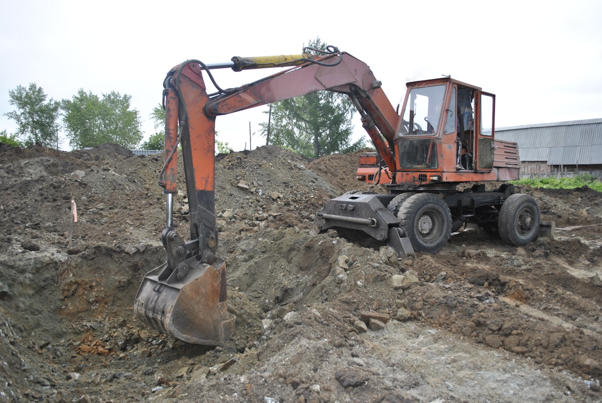 An old, rusty excavator is parked on rough terrain, surrounded by piles of dirt and gravel. The excavator appears to be in working condition with visible hydraulic components. It is set against a backdrop of trees and industrial buildings.
