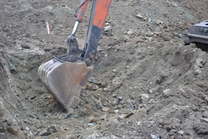 An excavator's bucket arm is actively digging into a rocky and gravel-filled ground. The metallic digging bucket is rusted and appears well-used, emphasizing the rough terrain it is working in. The soil is uneven with scattered rocks and debris visible throughout the excavation site.