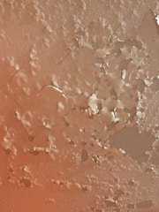 Peeling paint on a textured wall surface with various cracks and chipped areas. The paint has worn away in patches, revealing the layers beneath and creating an uneven, rugged appearance.