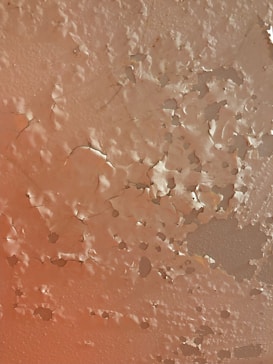 Peeling paint on a textured wall surface with various cracks and chipped areas. The paint has worn away in patches, revealing the layers beneath and creating an uneven, rugged appearance.