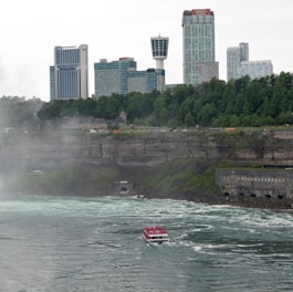 A red and white tourist boat navigates through swirling waters near a rocky shoreline. The background is dominated by a line of modern skyscrapers and a densely wooded area on a cliff. Mist rises from the churning water, adding a dynamic element to the scene.