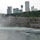 A red and white tourist boat navigates through swirling waters near a rocky shoreline. The background is dominated by a line of modern skyscrapers and a densely wooded area on a cliff. Mist rises from the churning water, adding a dynamic element to the scene.