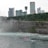A red and white tourist boat navigates through swirling waters near a rocky shoreline. The background is dominated by a line of modern skyscrapers and a densely wooded area on a cliff. Mist rises from the churning water, adding a dynamic element to the scene.