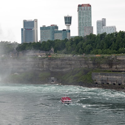 A red and white tourist boat navigates through swirling waters near a rocky shoreline. The background is dominated by a line of modern skyscrapers and a densely wooded area on a cliff. Mist rises from the churning water, adding a dynamic element to the scene.