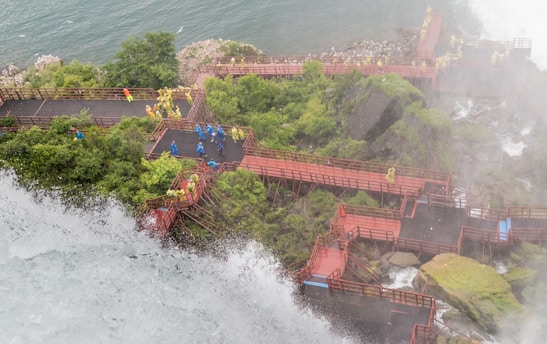 A series of red wooden walkways and platforms are constructed along a rocky, waterfall-laden landscape. People wearing yellow and blue raincoats are exploring the area, with mist rising from the waterfall. Lush greenery surrounds the paths, and the river is visible in the background.