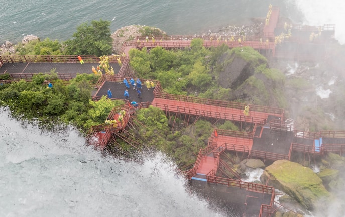 A series of red wooden walkways and platforms are constructed along a rocky, waterfall-laden landscape. People wearing yellow and blue raincoats are exploring the area, with mist rising from the waterfall. Lush greenery surrounds the paths, and the river is visible in the background.