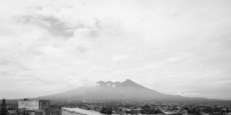 A black and white image featuring a wide landscape view with a prominent volcano in the distance. The foreground consists of urban elements with various buildings scattered throughout. The sky is filled with scattered clouds, contributing to a serene and expansive atmosphere.
