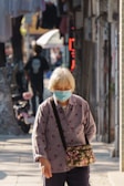An elderly person wearing a face mask walks down a narrow street, carrying a floral patterned bag. The street is lined with buildings, and there is another person walking in the background. Sunlight casts shadows, creating a warm atmosphere.
