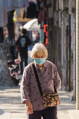 An elderly person wearing a face mask walks down a narrow street, carrying a floral patterned bag. The street is lined with buildings, and there is another person walking in the background. Sunlight casts shadows, creating a warm atmosphere.