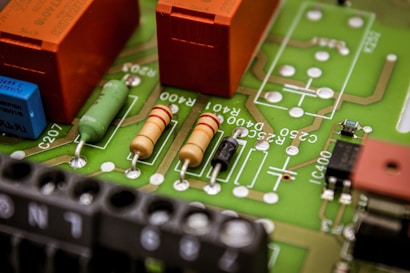 A close-up view of a printed circuit board featuring various electronic components such as resistors, capacitors, and connectors. The board has visible green solder mask and orange, black, and beige components mounted on it, with white markings for labeling.