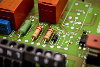 A close-up view of a printed circuit board featuring various electronic components such as resistors, capacitors, and connectors. The board has visible green solder mask and orange, black, and beige components mounted on it, with white markings for labeling.
