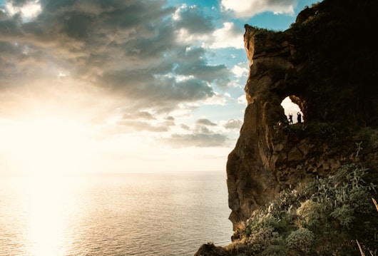 A dramatic seascape with a large rocky cliff on the right, featuring a natural arch formation in the rock. The sun is setting or rising, casting a warm glow over the ocean on the left. Clouds scatter across the blue sky, creating a picturesque scene. Several silhouetted figures stand inside the arch, adding a sense of scale.