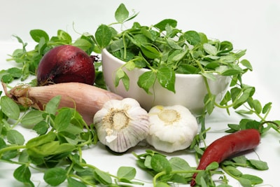 A white bowl filled with fresh green pea shoots surrounded by various vegetables. A red onion and a whole garlic bulb sit nearby, along with a shallot and a red chili pepper. The greens are vibrant, with droplets of water on some leaves, creating a fresh and organic feel.