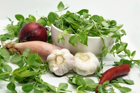 A white bowl filled with fresh green pea shoots surrounded by various vegetables. A red onion and a whole garlic bulb sit nearby, along with a shallot and a red chili pepper. The greens are vibrant, with droplets of water on some leaves, creating a fresh and organic feel.
