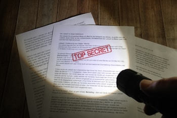 A hand holding a flashlight illuminates documents laid on a wooden surface. One document prominently displays a red 'TOP SECRET' stamp. The text is primarily in German, and the scene suggests a clandestine or investigative atmosphere.