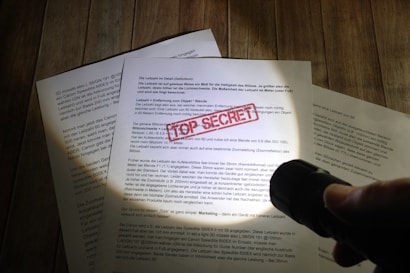 A hand holding a flashlight illuminates documents laid on a wooden surface. One document prominently displays a red 'TOP SECRET' stamp. The text is primarily in German, and the scene suggests a clandestine or investigative atmosphere.