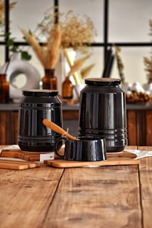 A rustic setting with two large black jars placed on wooden boards. A smaller black cup with a wooden-handled knife rests in front. The backdrop includes dried plants in amber glass bottles and a window with a grid pattern, creating a warm and cozy ambience.