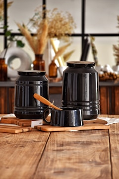 A rustic setting with two large black jars placed on wooden boards. A smaller black cup with a wooden-handled knife rests in front. The backdrop includes dried plants in amber glass bottles and a window with a grid pattern, creating a warm and cozy ambience.