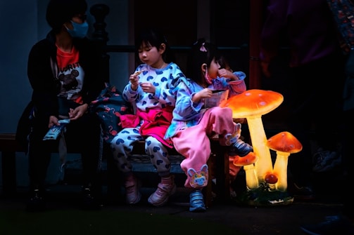 Two children wearing colorful clothing are seated on a bench. They are engaging with each other, possibly sharing food. A person beside them is wearing a mask and casual attire. Beside the children, there are large, glowing, fantasy-like mushroom decorations.