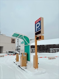 A snow-covered parking lot features a prominent parking sign with Japanese characters and English letters 'P' and 'IN'. The area includes automated parking fee machines and a green canopy. The lot is surrounded by industrial buildings and a few parked vehicles, with barriers and snow clearly visible.