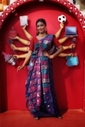 A woman in a colorful sari poses in front of a vibrant red backdrop featuring an illustration of multiple arms each holding different items like a newspaper, football, laptop, and microphone. The sari is predominantly blue with floral patterns in pink and green. She is adorned with traditional jewelry, including earrings and necklaces, and her expression is confident and poised.