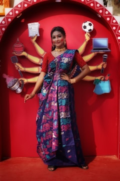 A woman in a colorful sari poses in front of a vibrant red backdrop featuring an illustration of multiple arms each holding different items like a newspaper, football, laptop, and microphone. The sari is predominantly blue with floral patterns in pink and green. She is adorned with traditional jewelry, including earrings and necklaces, and her expression is confident and poised.