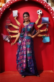 A woman in a colorful sari poses in front of a vibrant red backdrop featuring an illustration of multiple arms each holding different items like a newspaper, football, laptop, and microphone. The sari is predominantly blue with floral patterns in pink and green. She is adorned with traditional jewelry, including earrings and necklaces, and her expression is confident and poised.