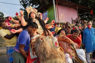 A group of people is carrying an elaborate statue of a multi-armed goddess, adorned with vibrant garments and jewelry. The goddess is depicted atop a lion, and several men are assisting in its movement. The background shows more people observing, some capturing the moment on their phones. A small structure is visible with lush greenery around.