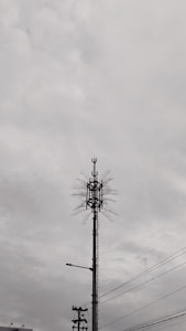 A tall communication tower with multiple antennas and cables is set against a cloudy sky. The tower features linear and dish components near the top, with surrounding power lines visible in the foreground.