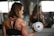 A woman with muscular arms lifts a dumbbell while facing a mirror. She is wearing a black strappy sports bra, and her focus is on her reflection. The background reveals a gym setting with various equipment visible.