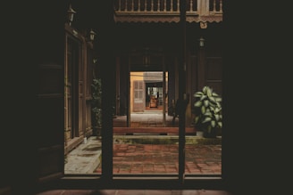 Sunlit courtyard of a traditional Kerala house with rustic wooden doors and lush tropical plants lining the stone pathway.