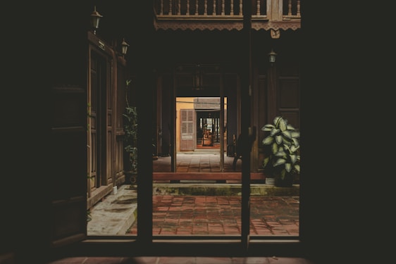 Sunlit courtyard of a traditional Kerala house with rustic wooden doors and lush tropical plants lining the stone pathway.