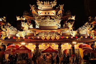 An ornate traditional Asian temple adorned with intricate designs and lit warmly at night. Colorful lanterns hang from the structure, with several red canopies in the foreground where people are gathered.