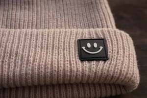 A close-up view of a knitted beige beanie featuring a black square patch with a white smiling face. The texture of the wool is clearly visible, and the image is set against a blurred dark background.